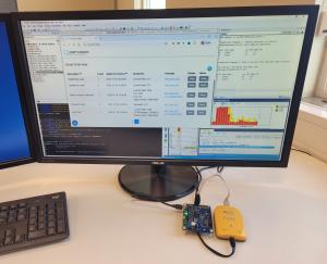 A computer workstation showing the Percepio Detect dashboard with example data from the GitHub demo displayed on a large monitor. The screen also shows IAR Embedded Workbench® windows, code editors, and diagnostic charts. On the desk below, a microcontrol