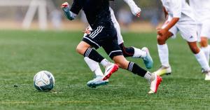 Youth soccer player dribbling a Futstrikers Tekno24 match ball in the rain during a competitive match while defenders chase in the background.