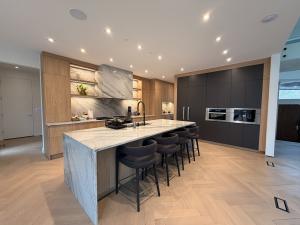 A wide-angle view of a luxury kitchen in Potomac, MD, showcasing the Neo LEAF Golden Oak series. A large kitchen island with a white-and-grey marble waterfall countertop and four black barstools sits at the center. The background features a full marble ba