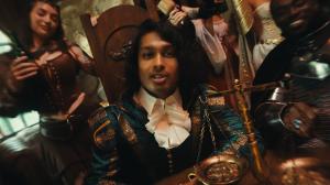 Ashwin Gane sits at a wooden table in elaborate teal and gold costume with a white ruffled shirt, surrounded by partygoers in fantasy outfits and a brass balance scale in the foreground.