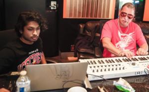 Ashwin Gane works on a laptop while Scott Storch plays a large MIDI keyboard beside him in a recording studio, surrounded by mixing desks and chairs.