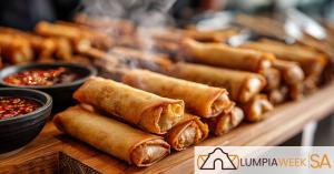 Close-up of golden-brown lumpiang shanghai on a wooden tray with dipping sauces and steam rising, with the Lumpia Week SA logo in the corner.