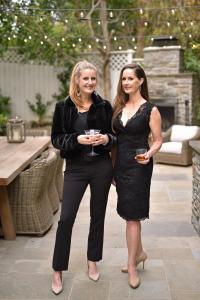 Two women in black dresses are holding cocktails and standing outside