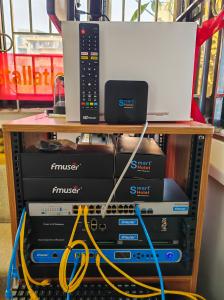 A physical demonstration rack of the FMUSER hotel IPTV system, showing the FBE013 set-top box, FBE 015 PLUS remote, network switch, Tuner to IP Gateway, and Multi-Channel CI-Receiver