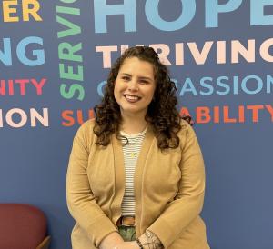 Allison Palmer, Chief Mission Officer of Horizon Goodwill, smiling with curly brown hair wearing a tan cardigan and striped shirt, standing in front of a blue wall with colorful words including 