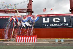 Balloons and streamers fall alongside the bulk carrier HSL Geraldton during a ceremonial celebration, with red-and-white decorations and U.S. and Japanese flags overhead.