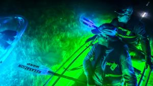 Two people in a transparent kayak with green LED glow, one holding a paddle labeled 