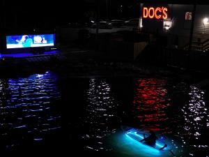 Nighttime photo of a large illuminated trailer screen playing a movie, reflecting on water with red and blue hues, a single blue-glowing kayak with paddler in foreground, and red neon 
