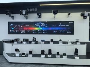 A clean photograph of a Hikvision showroom exhibit. The central wall display shows a colorful spectrum chart linking different wave types (sound, radio, micro, red-line, visible, purple, X-ray, Gamma) to specific camera technologies. Wall-mounted cameras 