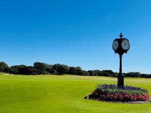 Freestanding black double-faced clock on a manicured green lawn, surrounded by flowers, with a wide golf course and clear blue sky in the background.