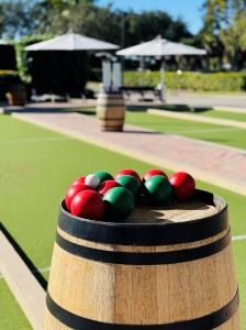 Close-up of red, green, and white bocce balls resting on a wooden barrel beside a bocce court, with umbrellas and seating blurred in the background.