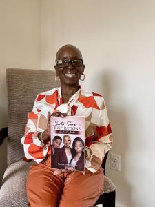 A portrait of a smiling Black woman with a shaved head and glasses, Tara Denise Tripp, sitting in a textured chair against a plain wall. She is holding up a copy of her new book, Sister Tara's Inspirations: Sister to Sister. She wears an orange and peach 