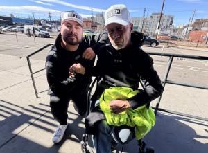 Dustin Folkes, founder of VYZN Apparel, giving clothing to a man in a wheelchair outdoors as part of the brand’s buy one gift one initiative supporting individuals experiencing homelessness.