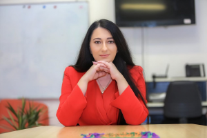 A professional portrait of Tammy Kassiou, Chair of Vorakai Lda. She has long dark hair and is wearing a vibrant red blouse, sitting at a wooden table with her hands clasped under her chin. The background shows a professional office setting with a whiteboa
