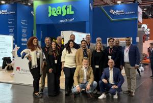 The Brazilian delegation poses in front of the Brazil stand at Interpack, inside an international trade fair pavilion. The group brings together professionals in business attire, with some standing and two crouched in front. In the background, the stand’s
