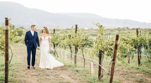 Bride and groom walking hand in hand through vineyard rows at Alcantara Vineyard wedding venue in Camp Verde, Arizona, with mountain views in the background.