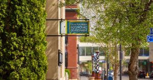 The Grand Forks Region Economic Development Corporation sign hanging from a building exterior on a tree-lined downtown street on a sunny spring day.