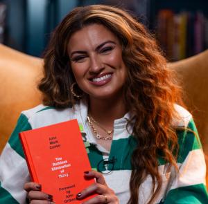 Tori Kelly sits in a high-backed leather chair, smiling while holding a bright orange book.
