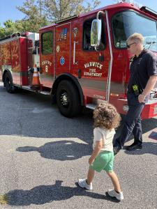 A young child with curly brown hair wearing a white t-shirt and green shorts walks across a sunny parking lot toward a large red Fire Department fire truck labeled Engine 8. A firefighter in a dark uniform stands near the truck watching the child approach