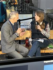 Portland Mayor Keith Wilson, holding a microphone, listens as Erin Holcomb of Portland Rescue Mission speaks beside him on a wooden bench along a city sidewalk.
