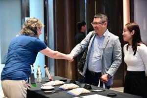 A woman shakes the hand of a smiling man at the 2026 U.S. Identity Preserved International Summit while another woman looks on.