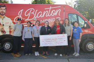 A J. Blanton Plumbing team member and six Orphans of the Storm staff members smile in front of a red J. Blanton Plumbing van, holding an oversized $5,000 check made out to Orphans of the Storm with the memo “Dogs Donation.”