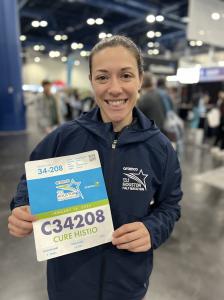 Medium height brown haired woman holding up a bib with her registration number and information for the Aramco Half-Marathon