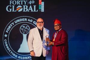 Ayodeji Razaq, dressed in a traditional burgundy agbada and red cap, receives the Forty Under 40 Global Award for Marketing & Communications 2025 on stage in Dubai, standing beside Mike Tankel in a white blazer. The event backdrop displays the award logo 