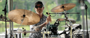A drummer passionately playing on stage, surrounded by cymbals and a drum set, with a natural landscape in the background.