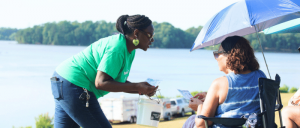 A woman in a green shirt engages with another person seated under an umbrella by a serene lakeside.