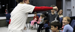 A person in a white shirt with red sleeves sprinkles a white substance over a child seated in a group of kids in a cozy indoor setting.