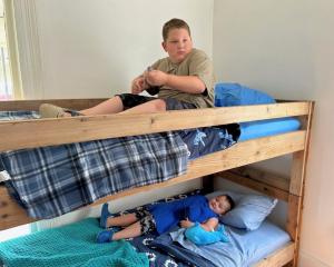 wo children enjoying their new wooden bunk bed from Sleep in Heavenly Peace. An older boy in a tan shirt sits cross-legged on the top bunk with blue bedding and a plaid blanket, while a younger child in a blue shirt lies on the bottom bunk with light blue
