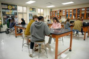 Students seated in groups in a GSSM physics classroom work on lab activities with a teacher assisting at the front of the room.